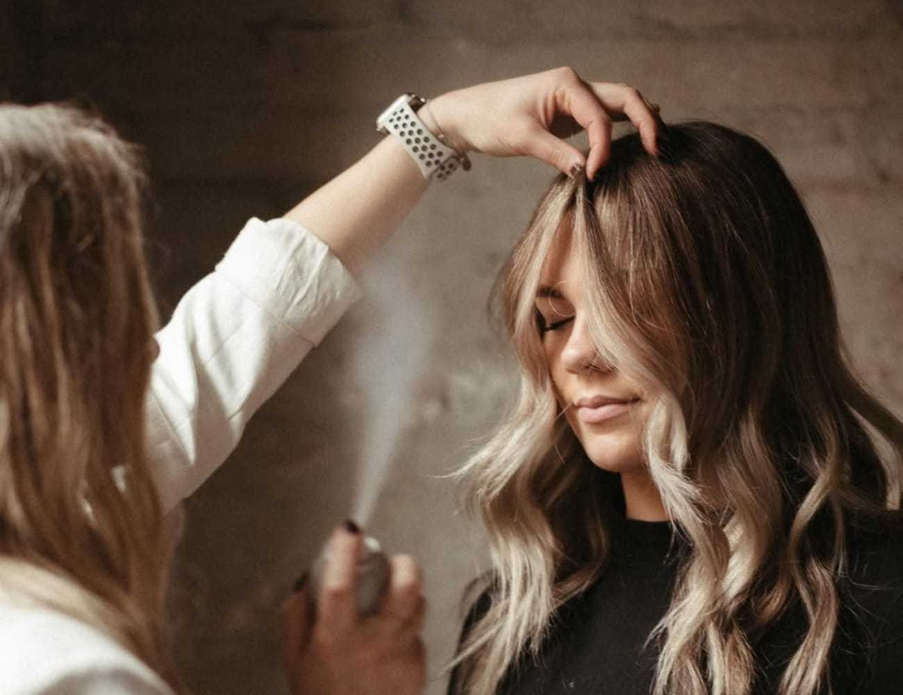 Hairstylist applying spray to woman with styled wavy hair in a salon setting.