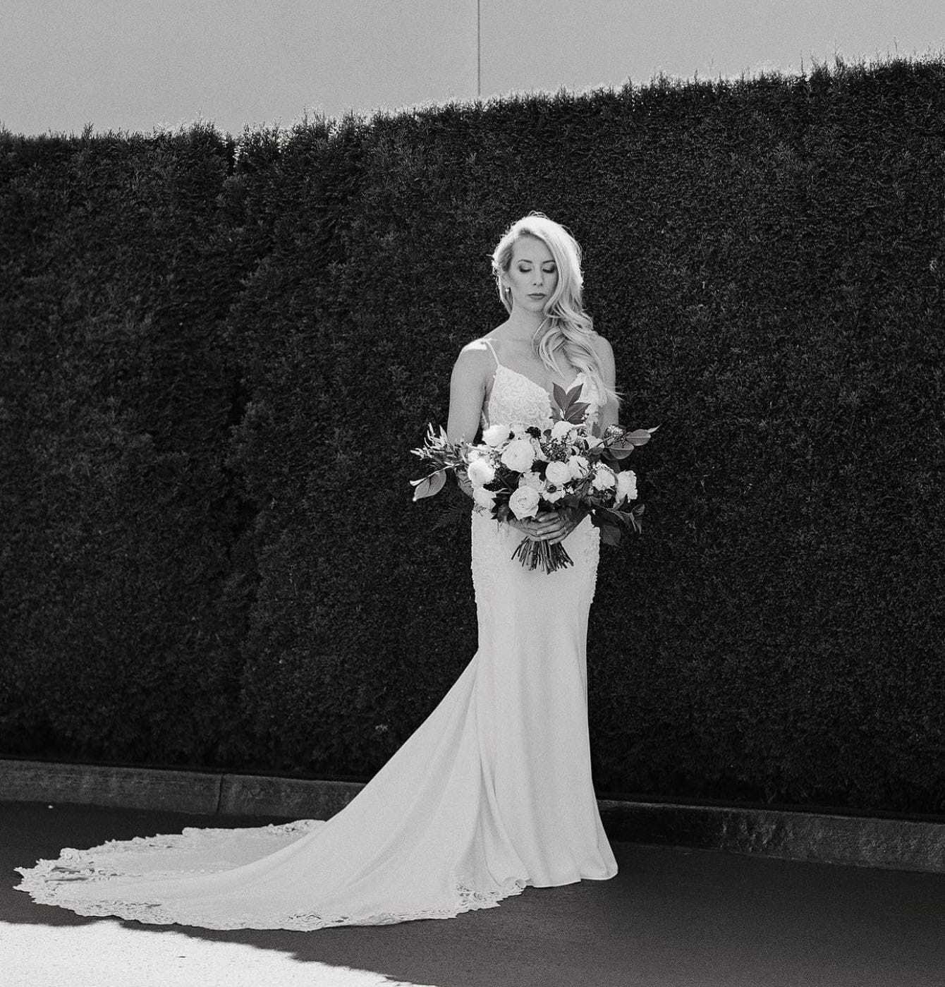 Bride in elegant gown holding a bouquet, standing by a manicured hedge.