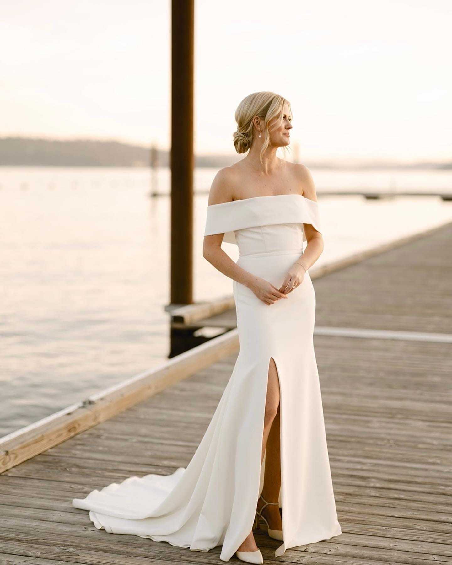 Bride in an off-shoulder gown stands on a dock, overlooking a serene water scene at sunset.