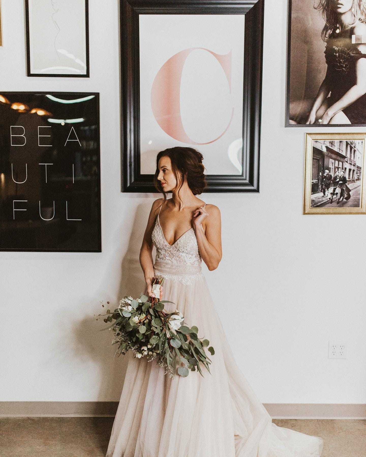 Bride in elegant gown holding bouquet, standing by framed wall art in stylish room.