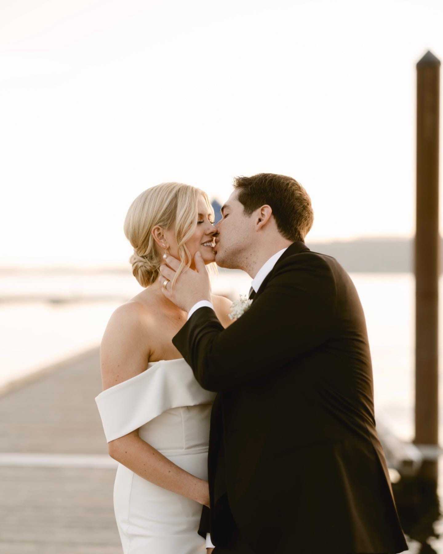Couple kisses on a dock at sunset, bride in off-shoulder gown, groom in black suit.